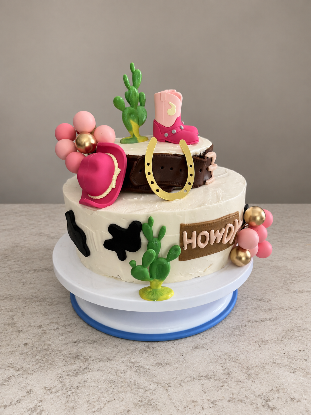 Two-tier country cake with pink cowboy accessories, cacti, cow spots, and a gold horseshoe